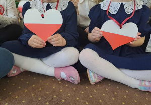 Dziewczynki w granatowych sukienkach trzymające biało-czerwone serca.