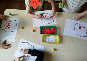 Dzieci wyklejają białymi i czerwonymi papierowymi kwiatkami szablon mapy Polski.